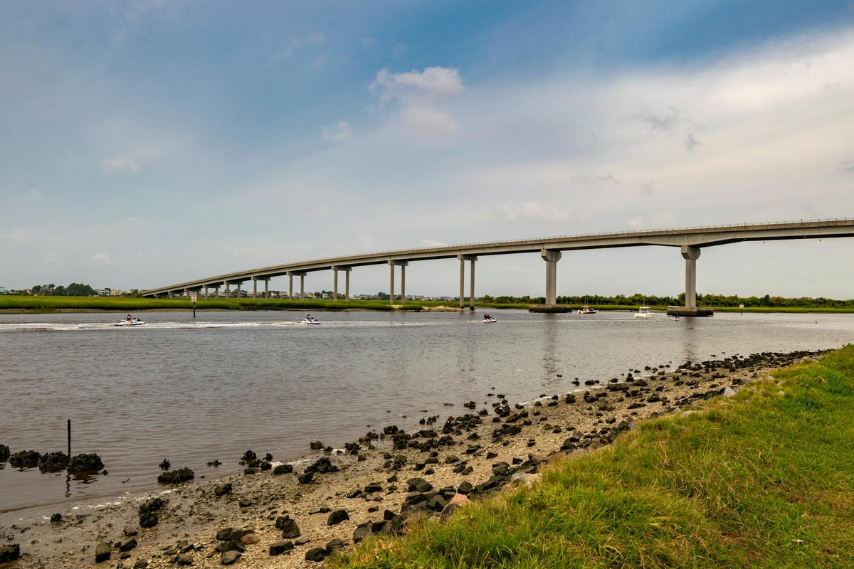 A long bridge spans over a calm river, surrounded by lush greenery under a blue sky.