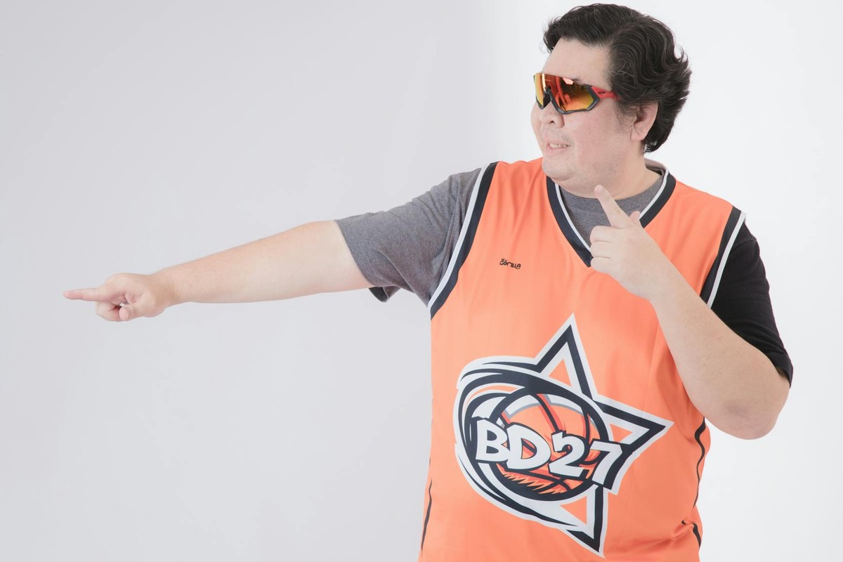 Asian man in orange sports jersey posing in studio with sunglasses.