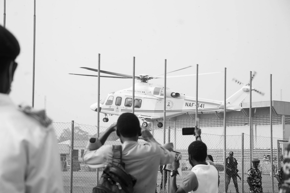 Black and white image of a military helicopter taking off, observed by people behind a fence.