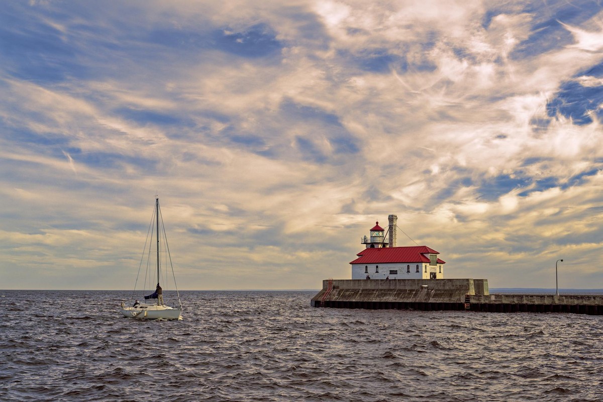Duluth lighthouse with sailboat on a serene day under a dramatic sky.