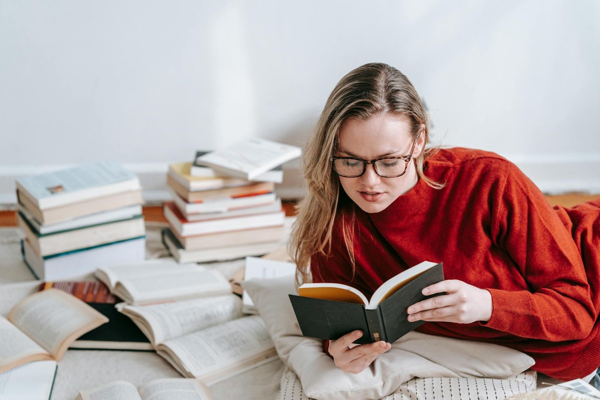 Intelligent female in eyeglasses reading book while lying on on floor with cushion and pile of literature in light room