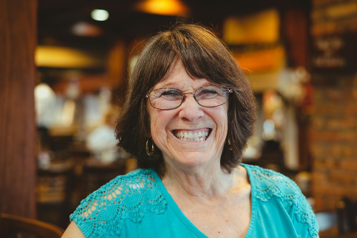 Cheerful elderly woman with glasses smiling brightly in a warm indoor environment.