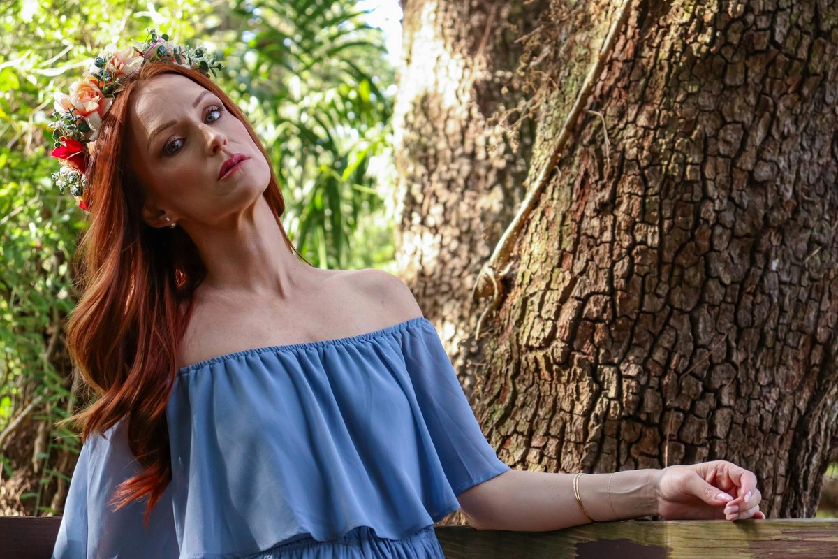 Woman wearing a blue dress and flower crown posing outdoors in a sunny park or forest setting.