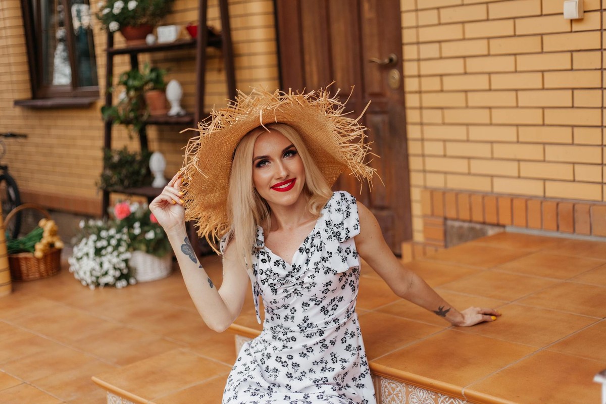 A cheerful woman wearing a straw hat and white sundress, sitting on steps outdoors with floral background.