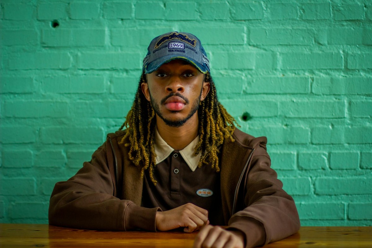 Stylish man in a brown jacket with dreadlocks and a cap sitting by a green brick wall.