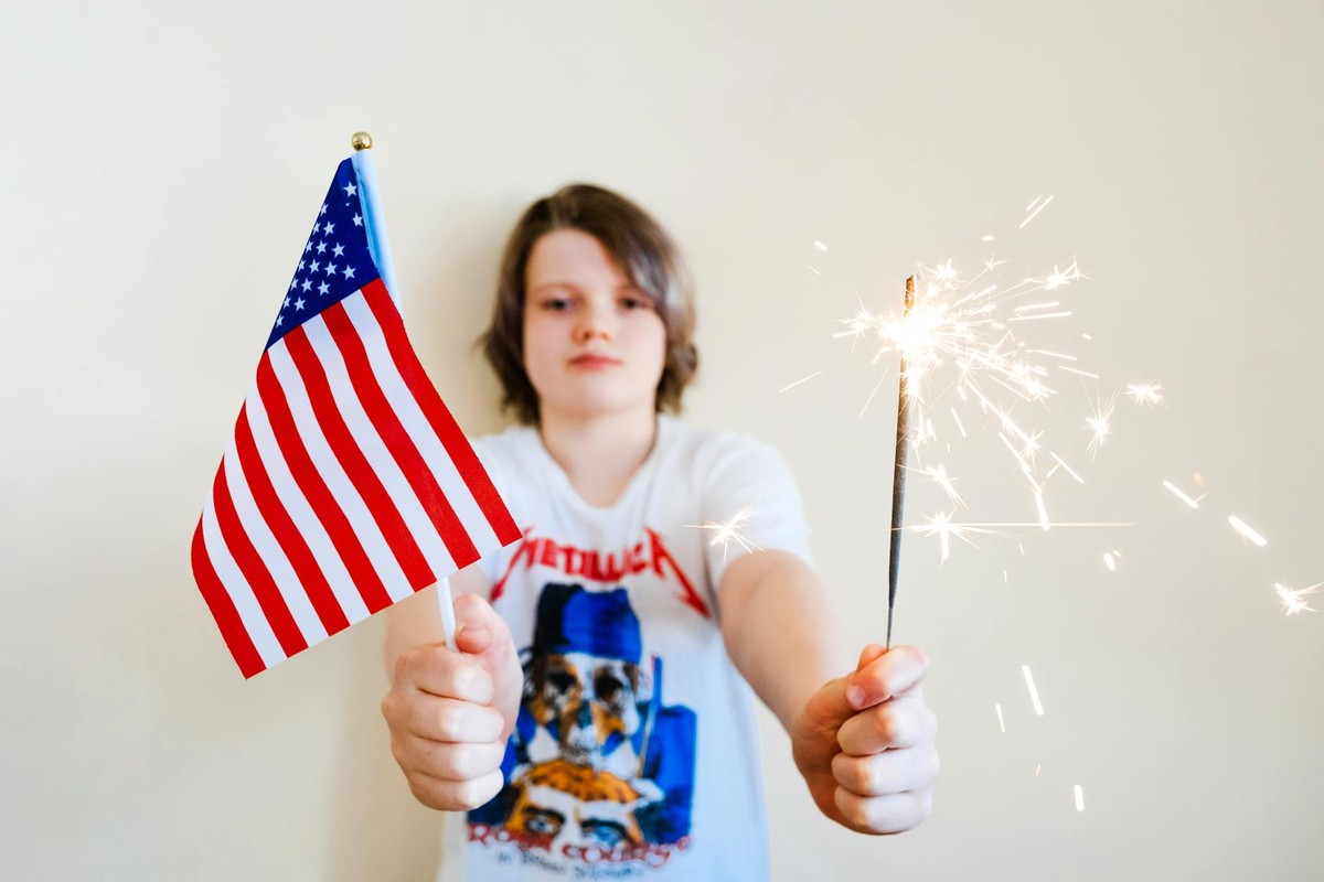 Young child joyfully holding American flag and sparkler indoors, celebrating USA holidays.