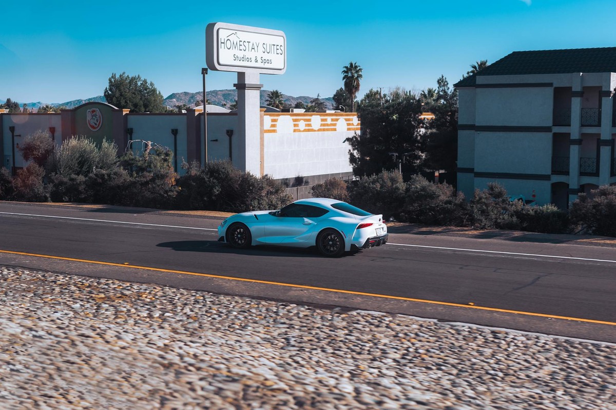 Blue Toyota GR Supra driving on road past Homestay Suites. Sunny day, clear sky.