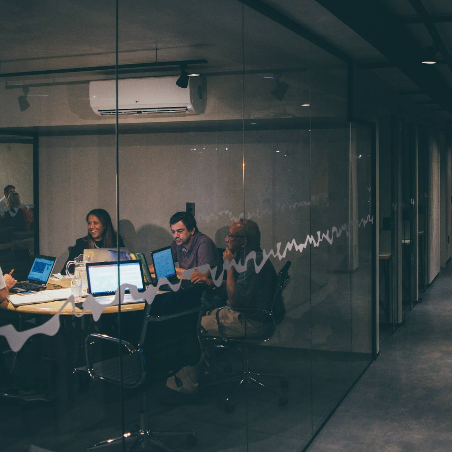 Group of four people engaged in a meeting around a table with laptops in a glass-walled office room.