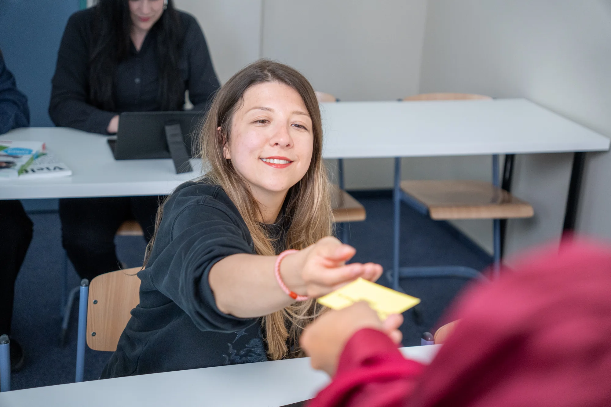 Lächelnde Frau reicht im Klassenzimmer eine gelbe Karte an eine andere Person.