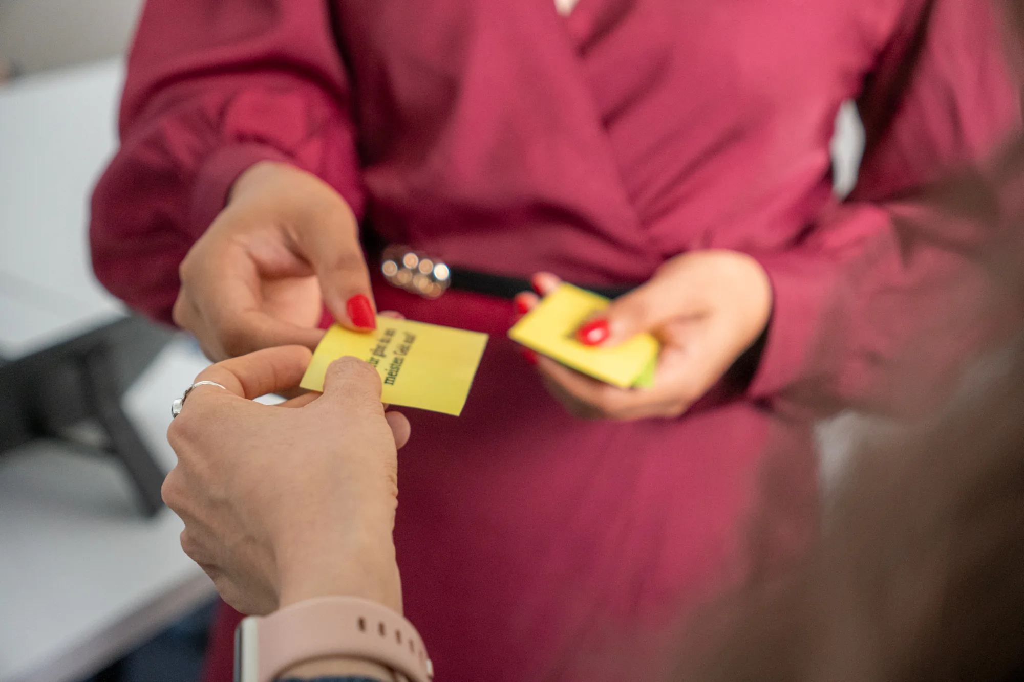 Eine Frau in rotem Kleid übergibt einer anderen Person gelbe Notizzettel mit Text.