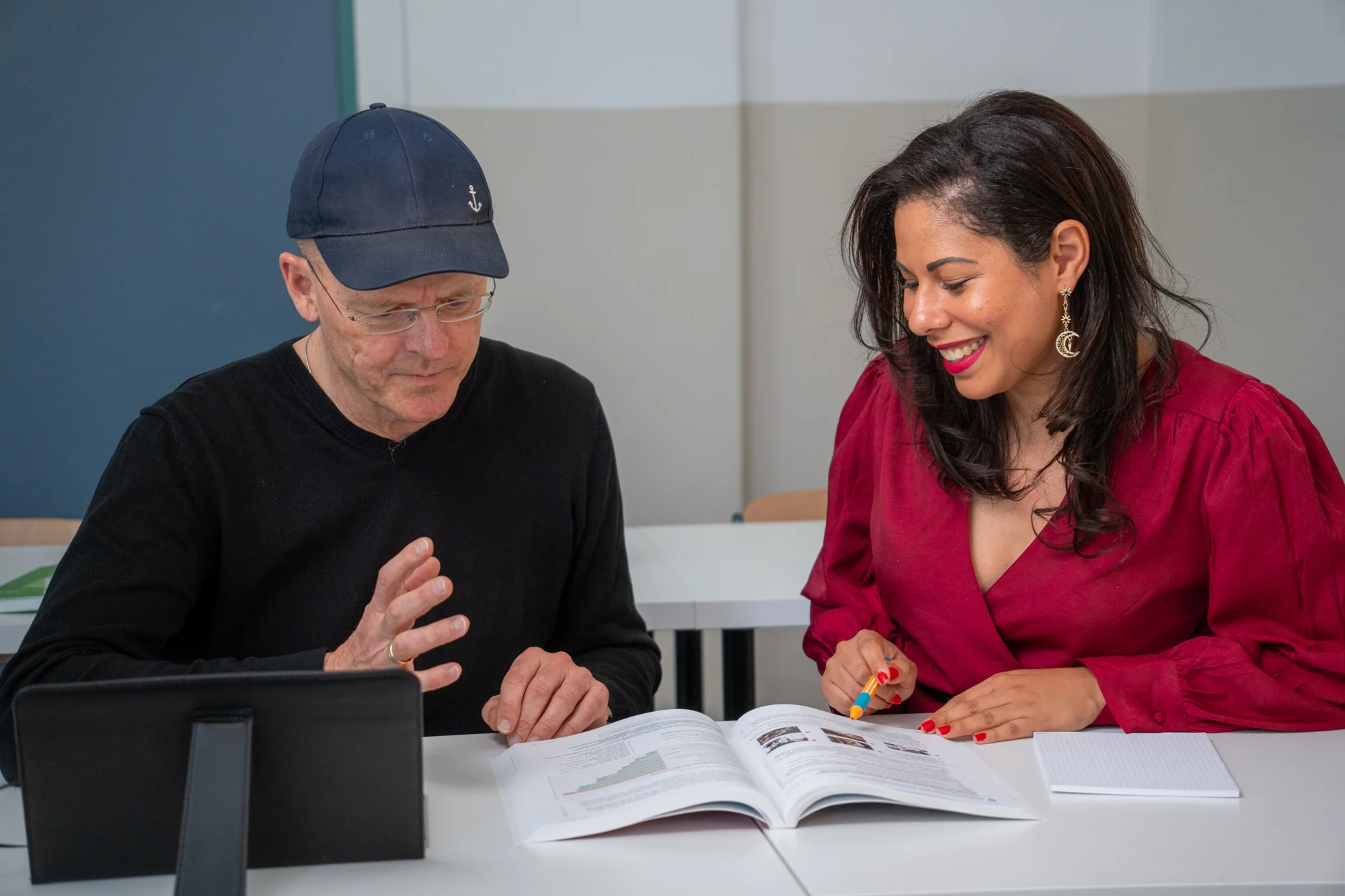 Ein Mann mit schwarzer Kappe und Frau in rotem Oberteil lesen gemeinsam ein Buch am Tisch.