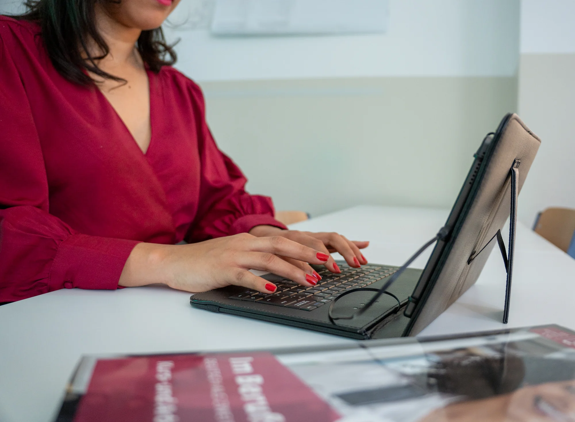 Frau in roter Bluse tippt auf einer Tablet-Tastatur an einem weißen Tisch.