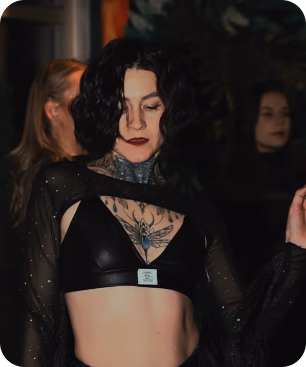 Woman with curly black hair, dark lipstick, and visible chest tattoos wearing a black crop top with sheer sleeves.