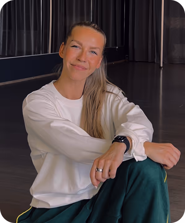 Smiling woman with long hair sitting on floor wearing a white sweatshirt and dark green pants in a room with dark curtains.