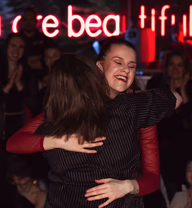 Woman with curly dark hair, a prominent chest tattoo, and dark lipstick wearing a black top in a dimly lit setting.