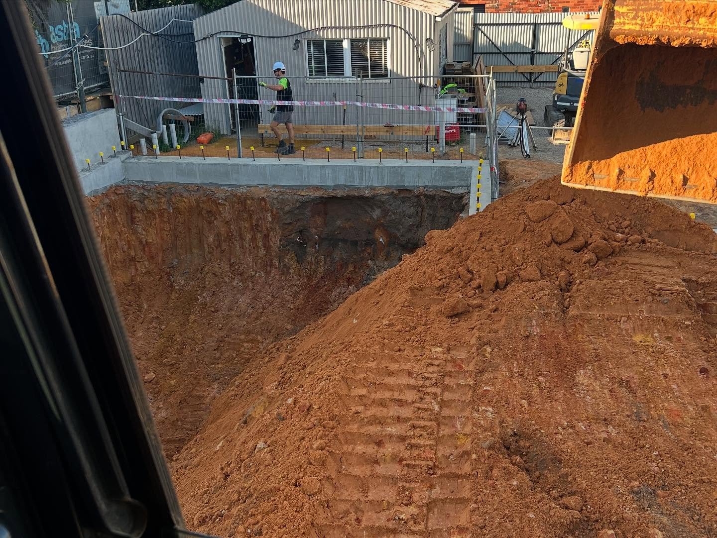 Construction site with an excavator moving a large pile of reddish soil into a deep rectangular excavation, and a worker in safety gear near the perimeter.