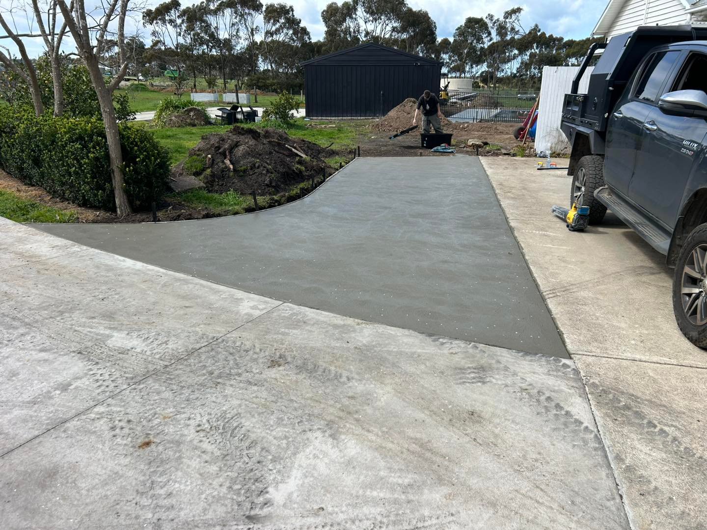 Freshly poured concrete driveway section in a residential outdoor area with a person working near a black shed and a parked dark pickup truck.