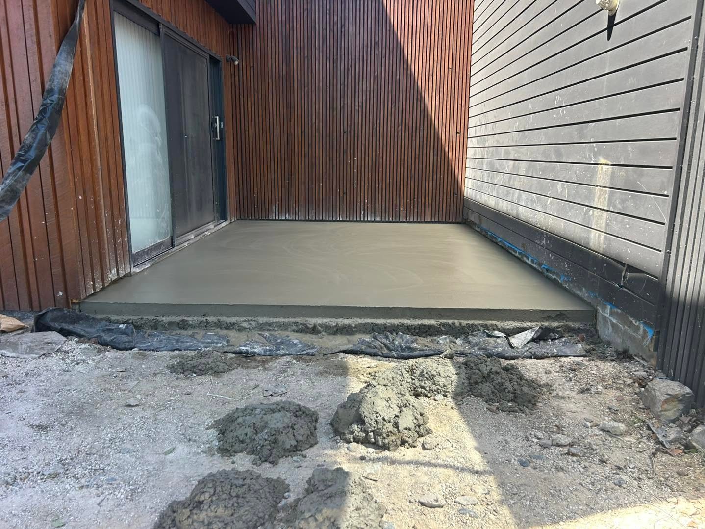 Newly poured smooth concrete patio slab between wooden vertical slat and horizontal siding walls with dirt and cement piles in front.