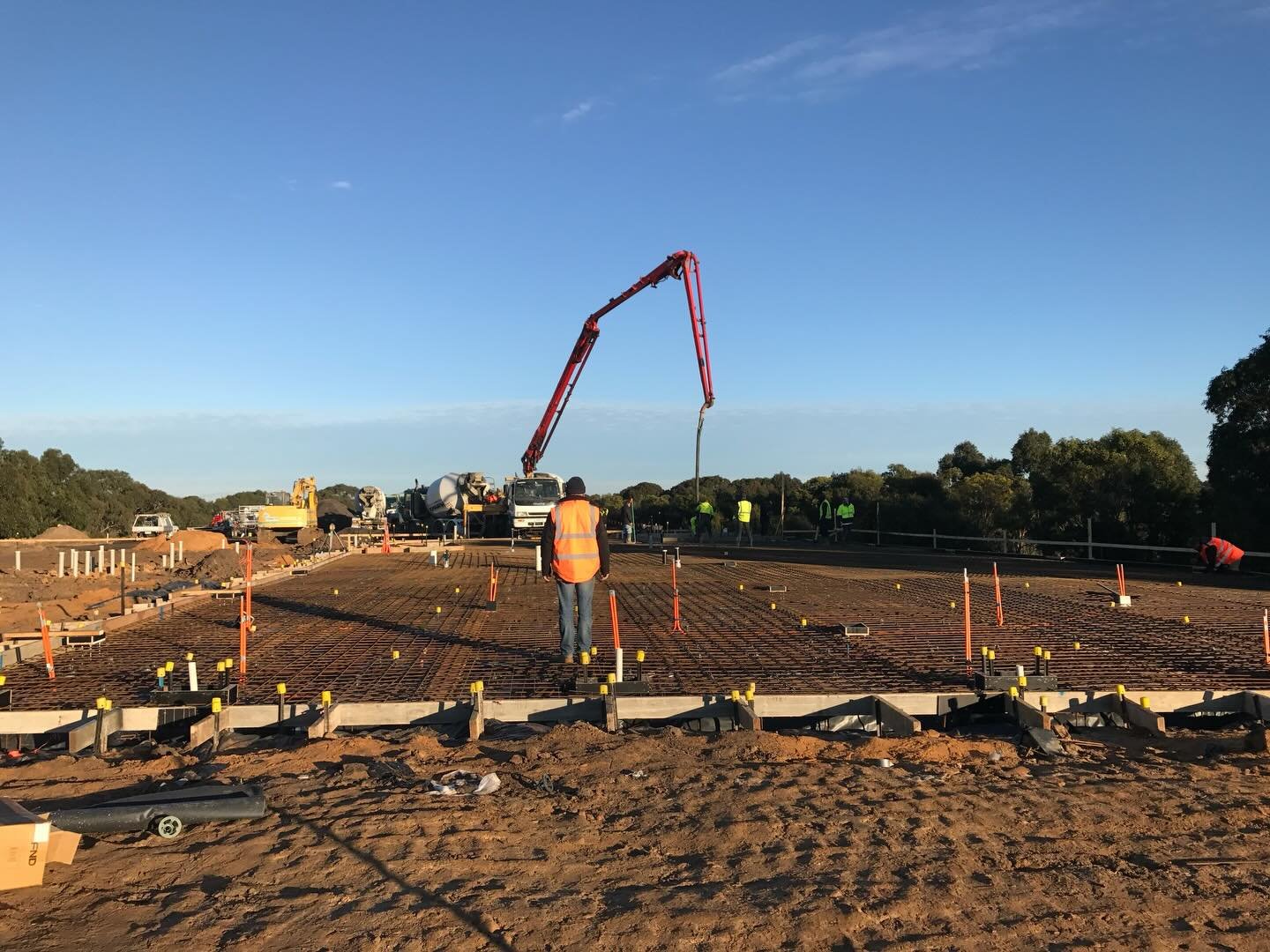 Construction workers wearing safety vests and equipment working on a concrete foundation with rebar, with a concrete pump truck in the background under a clear blue sky.