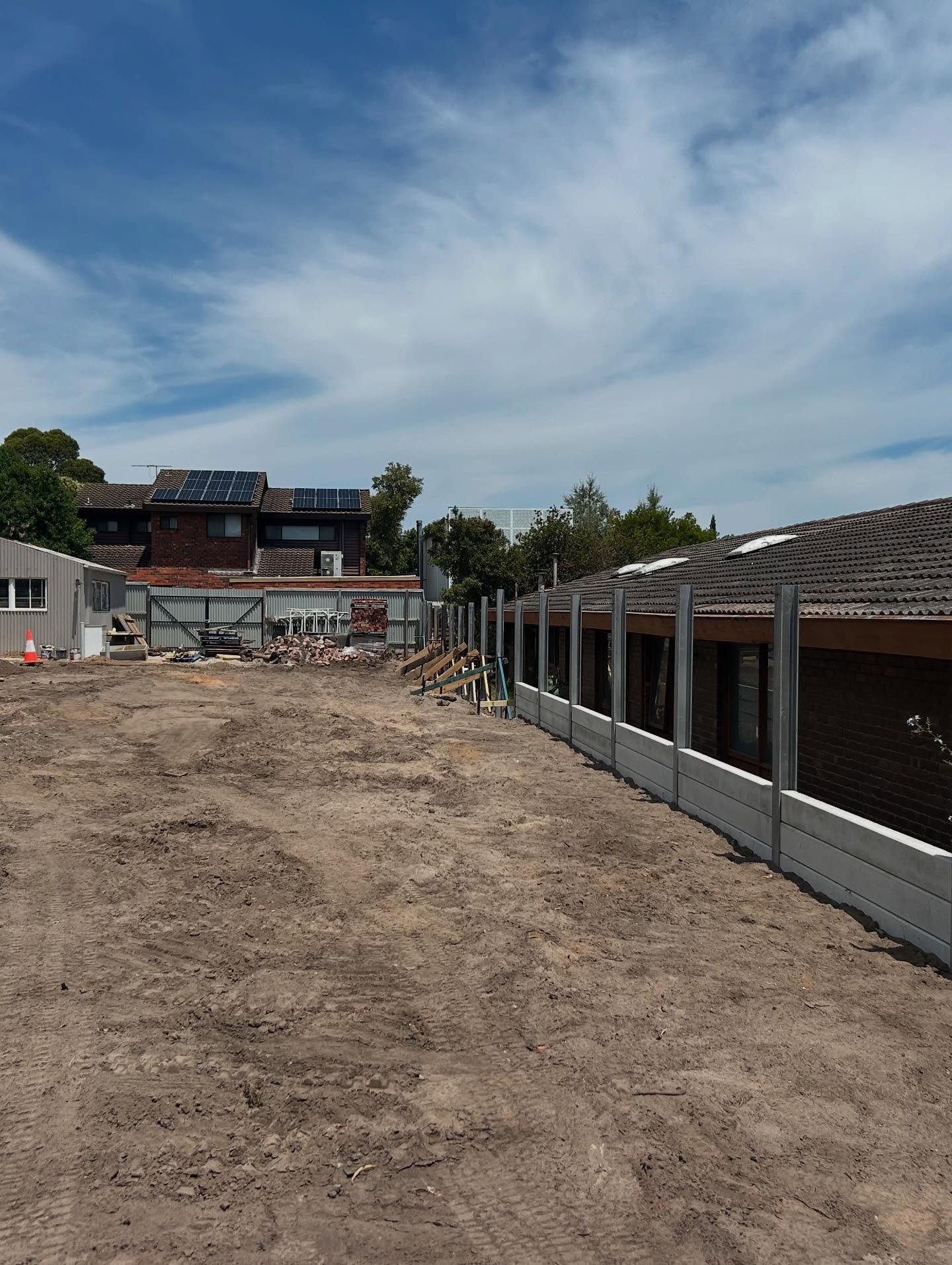 Construction site with cleared dirt ground and partially built concrete and metal fence next to a tiled roof building under a partly cloudy sky.