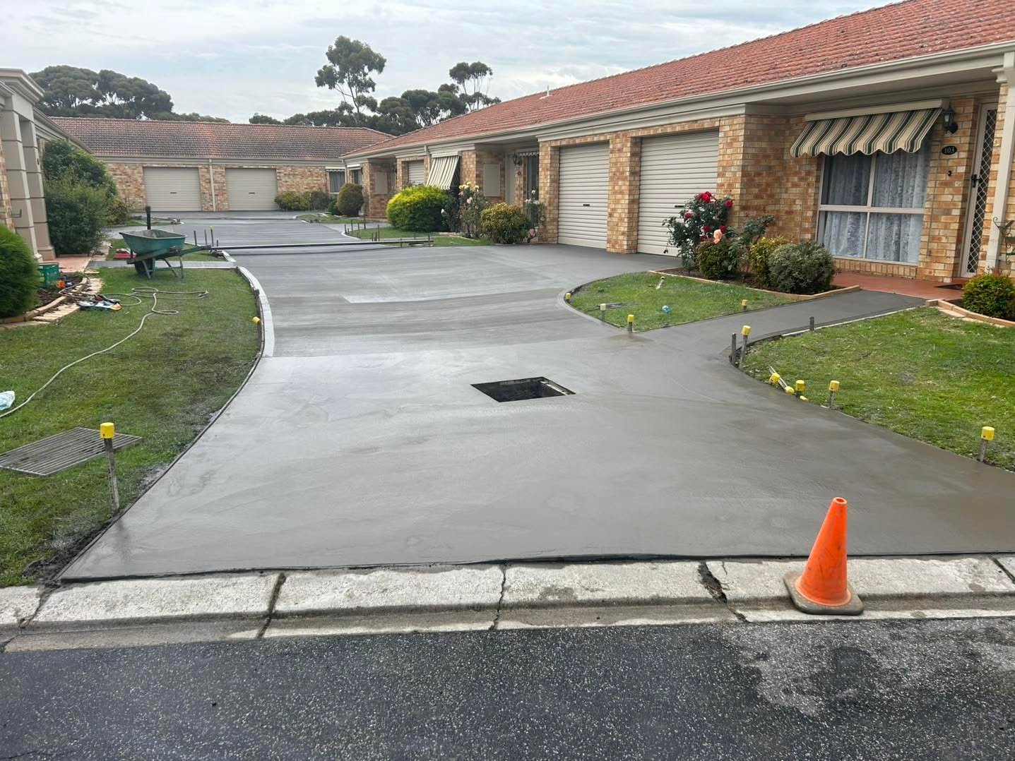 Freshly poured concrete driveway in front of brick residential garages, bordered by grass and marked with a single orange traffic cone.
