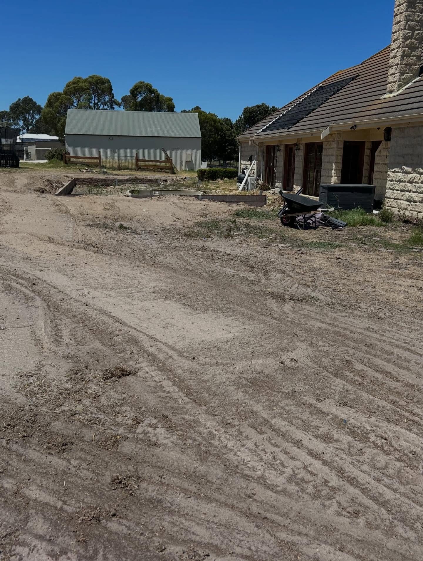 Construction site with a dirt ground showing tire tracks, a beige stone house on the right, and a metal barn in the background under a clear blue sky.