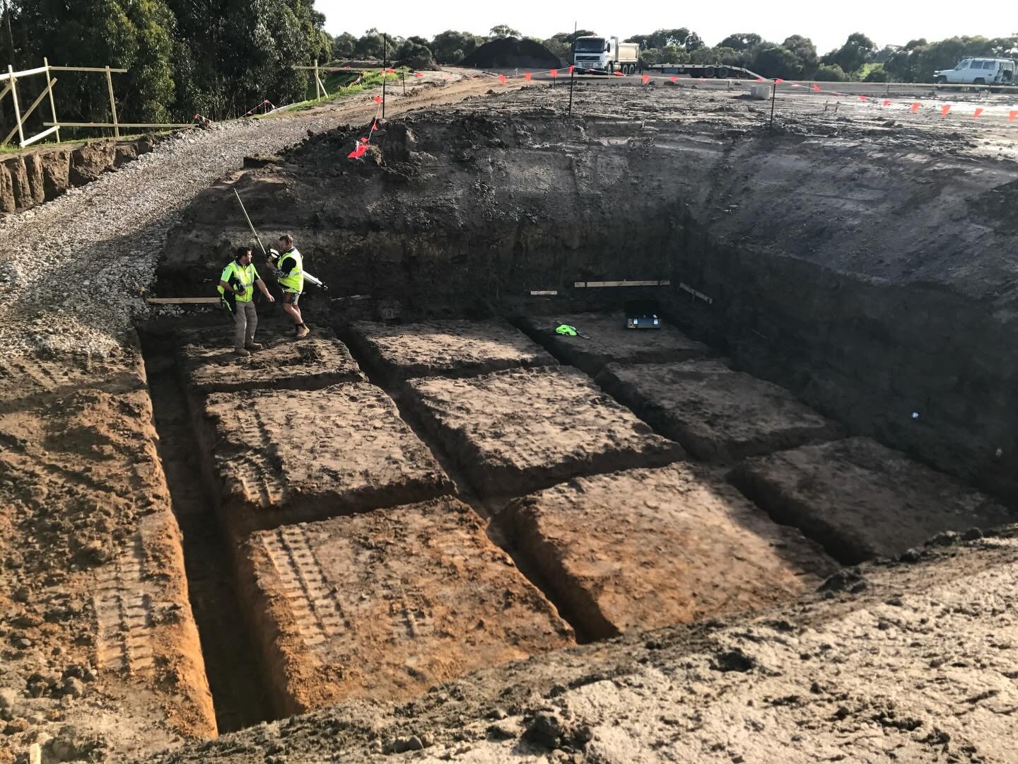 Two construction workers in high-visibility clothing standing inside a large segmented excavation pit on a construction site with vehicles and trees in the background.