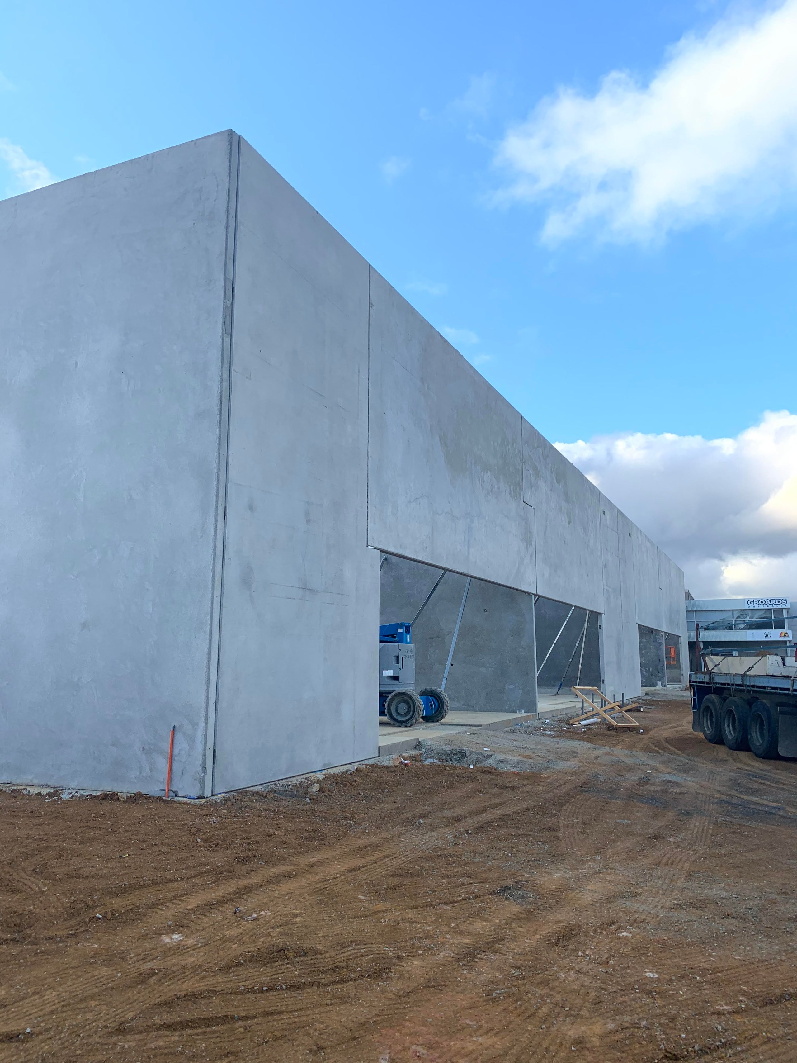 Under-construction concrete building with large open sections and construction equipment on a dirt ground under a blue sky.