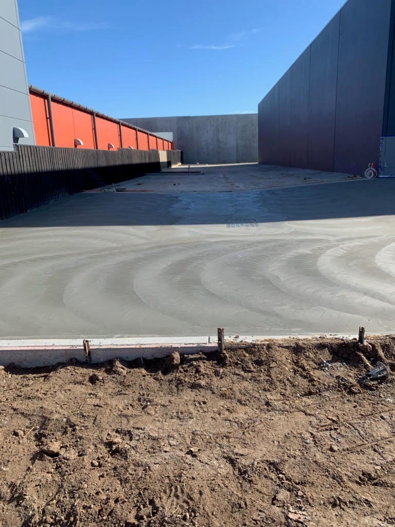 Freshly poured and smoothed concrete slab between industrial buildings under clear blue sky.