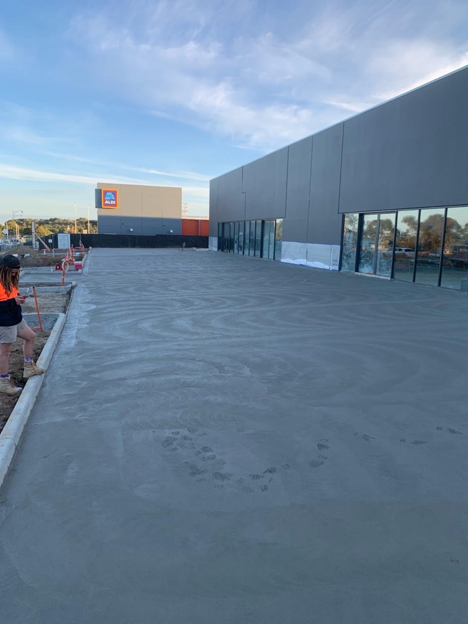 Newly poured concrete slab outside a commercial building with glass windows and an Aldi store visible in the background under a partly cloudy sky.