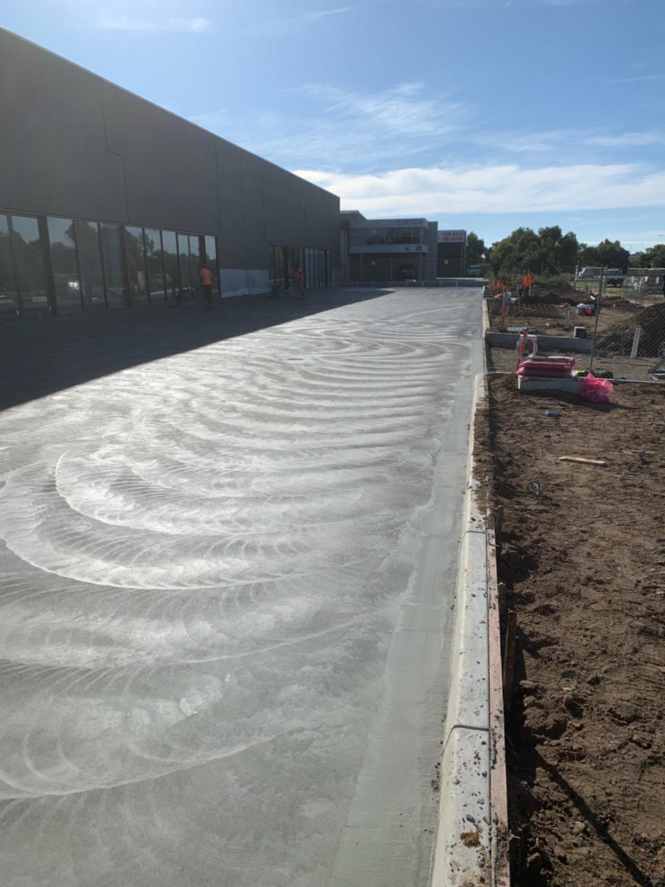 Newly poured and smoothed concrete pavement beside a large commercial building under a blue sky.