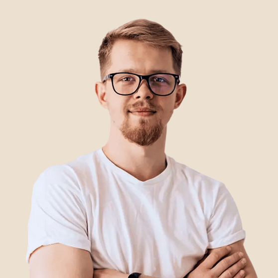 Young man with light brown hair, beard, and glasses wearing a white t-shirt with arms crossed against beige background.