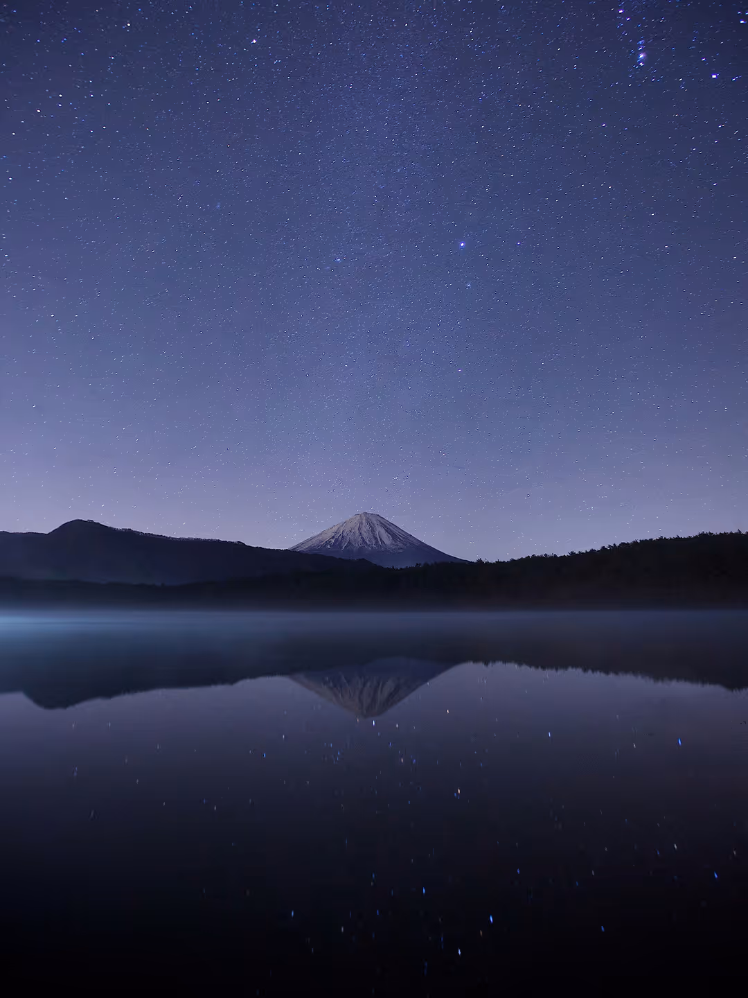 Snow-capped mountain under a starry night sky reflected in calm water.