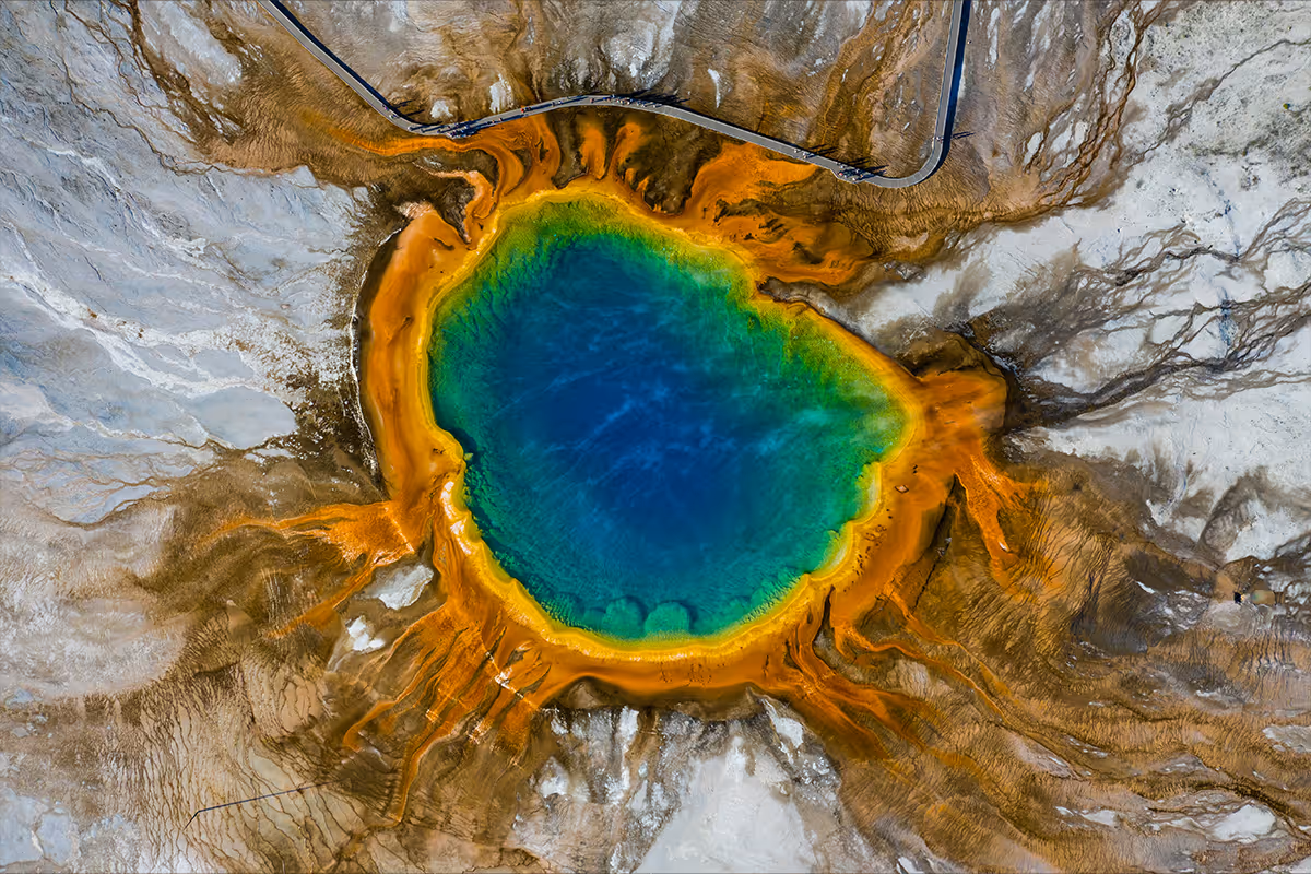 Aerial view of a colorful geothermal hot spring with blue center and orange bacterial mats around it.