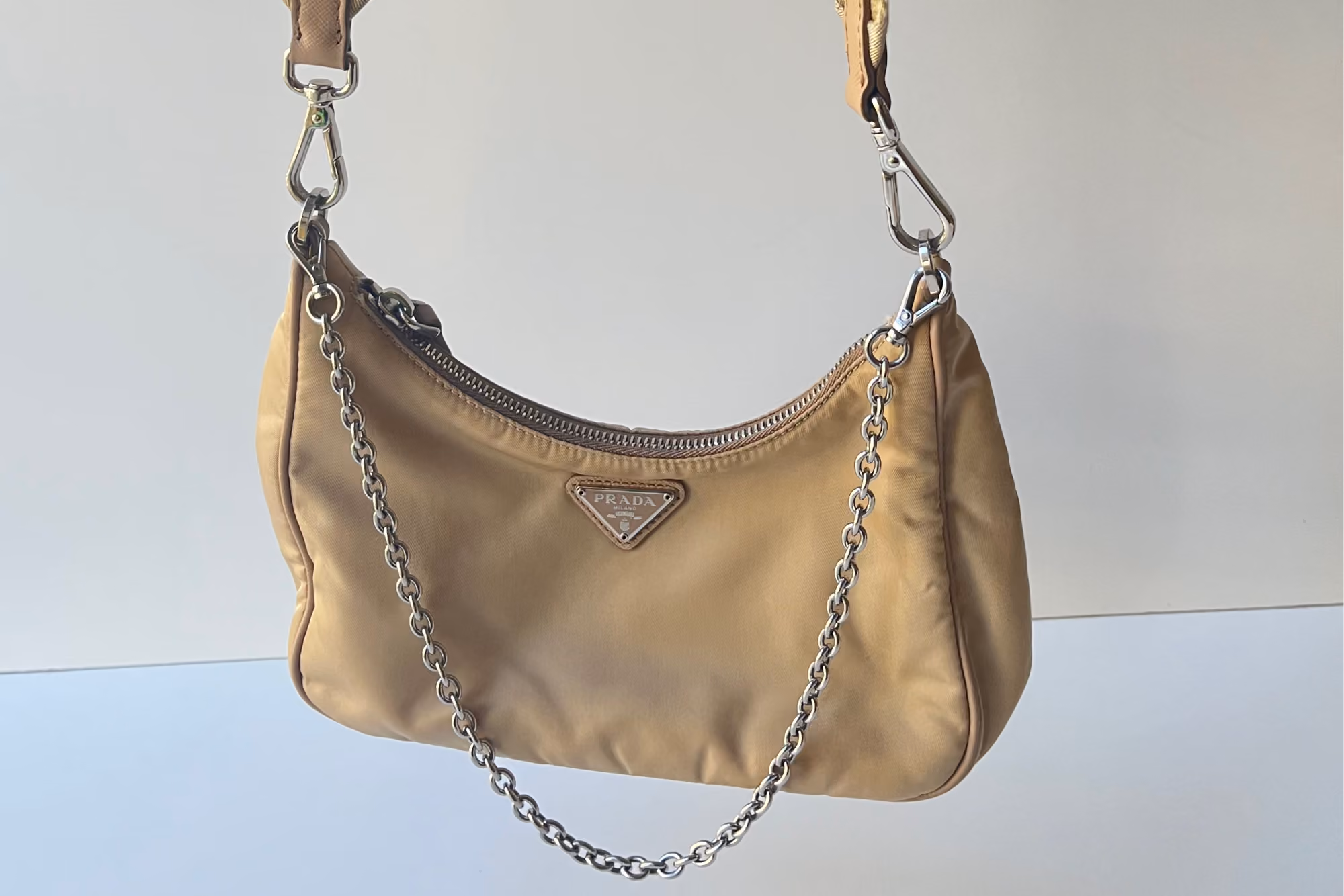 Beige nylon Prada Re-Edition hobo bag with triangular logo plaque and silver chain strap, hanging against a plain background.