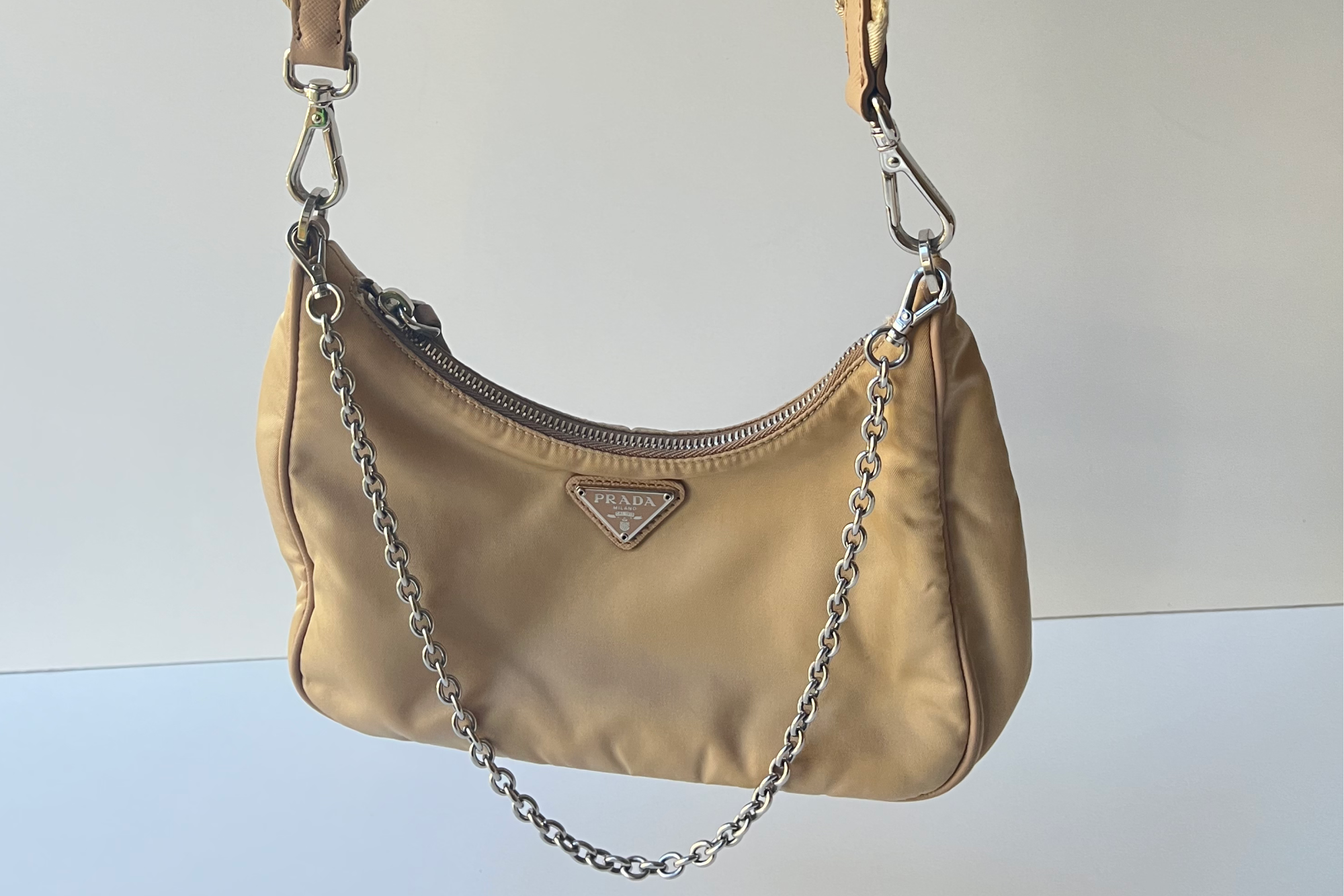 Beige nylon Prada Re-Edition hobo bag with triangular logo plaque and silver chain strap, hanging against a plain background.