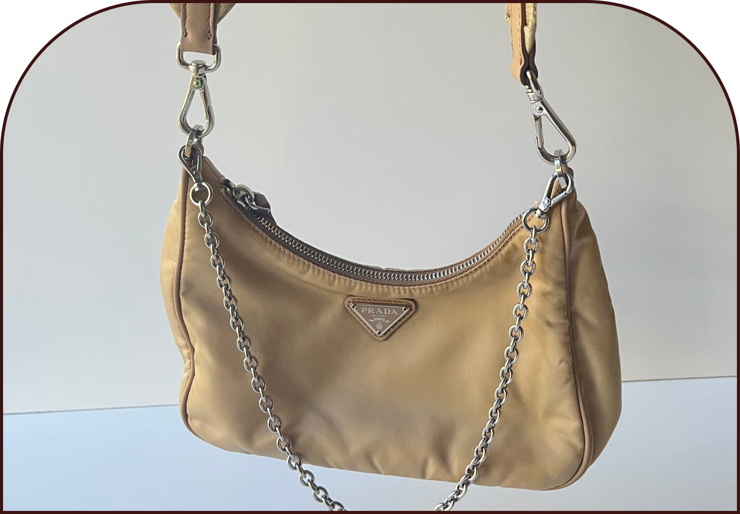 Beige nylon Prada Re-Edition hobo bag with triangular logo plaque and silver chain strap, hanging against a plain background.