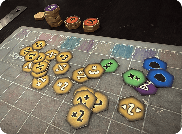 Prototype hexagonal game tokens and colored essence pieces arranged on a cutting mat.