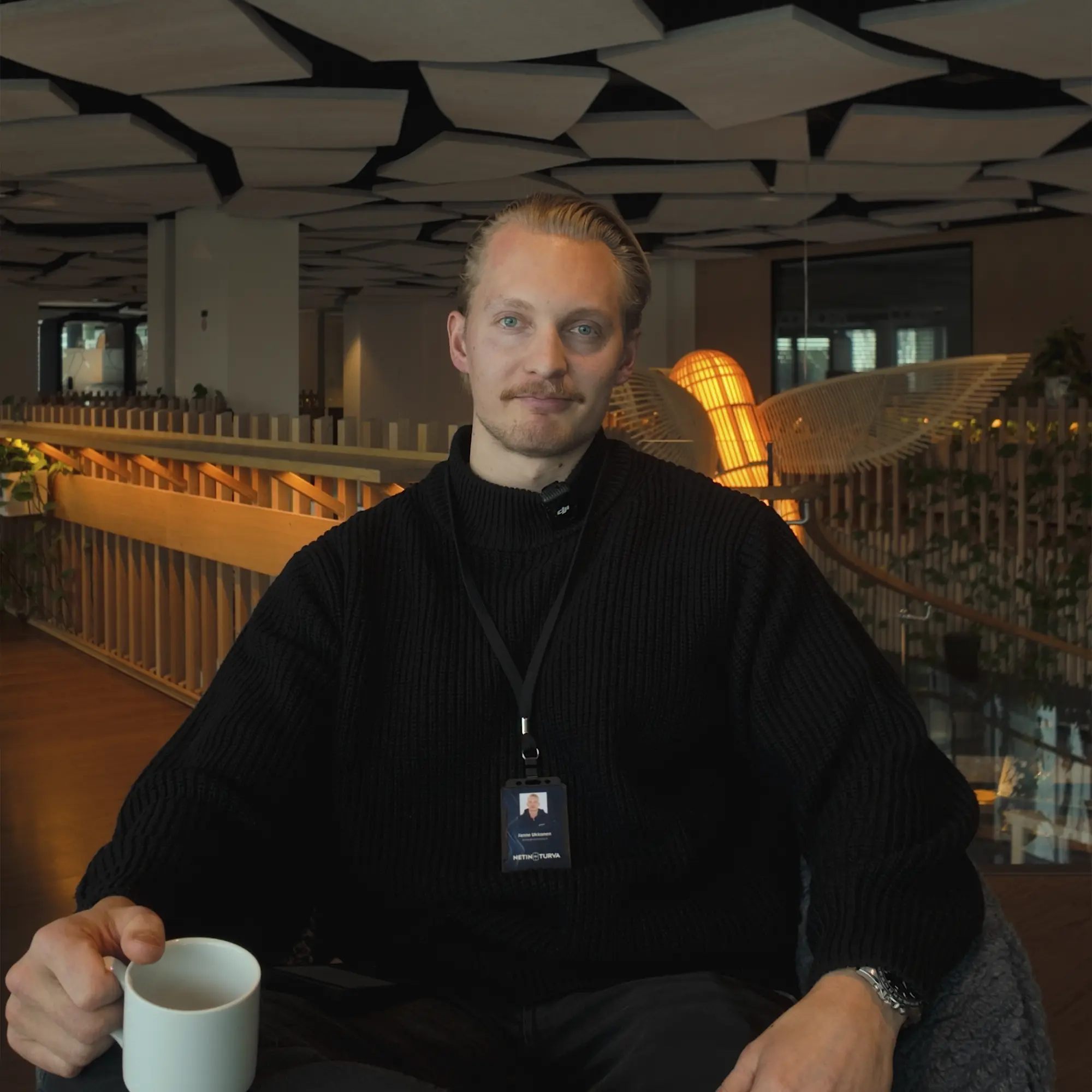 Man in black sweater holding a white mug seated in a modern room with wooden and light decor.