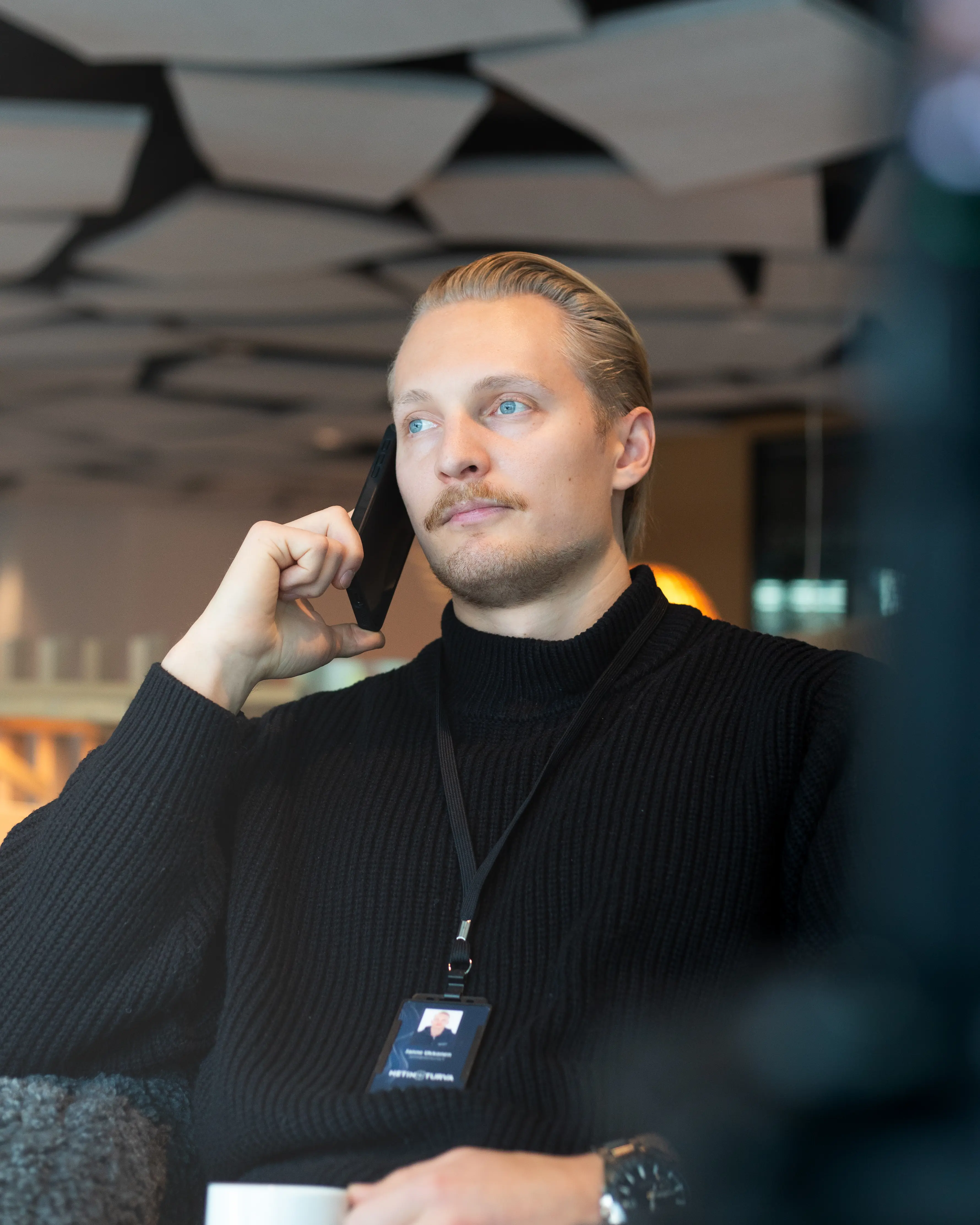 Blond man in black sweater holding phone to ear, wearing lanyard with badge, sitting indoors.