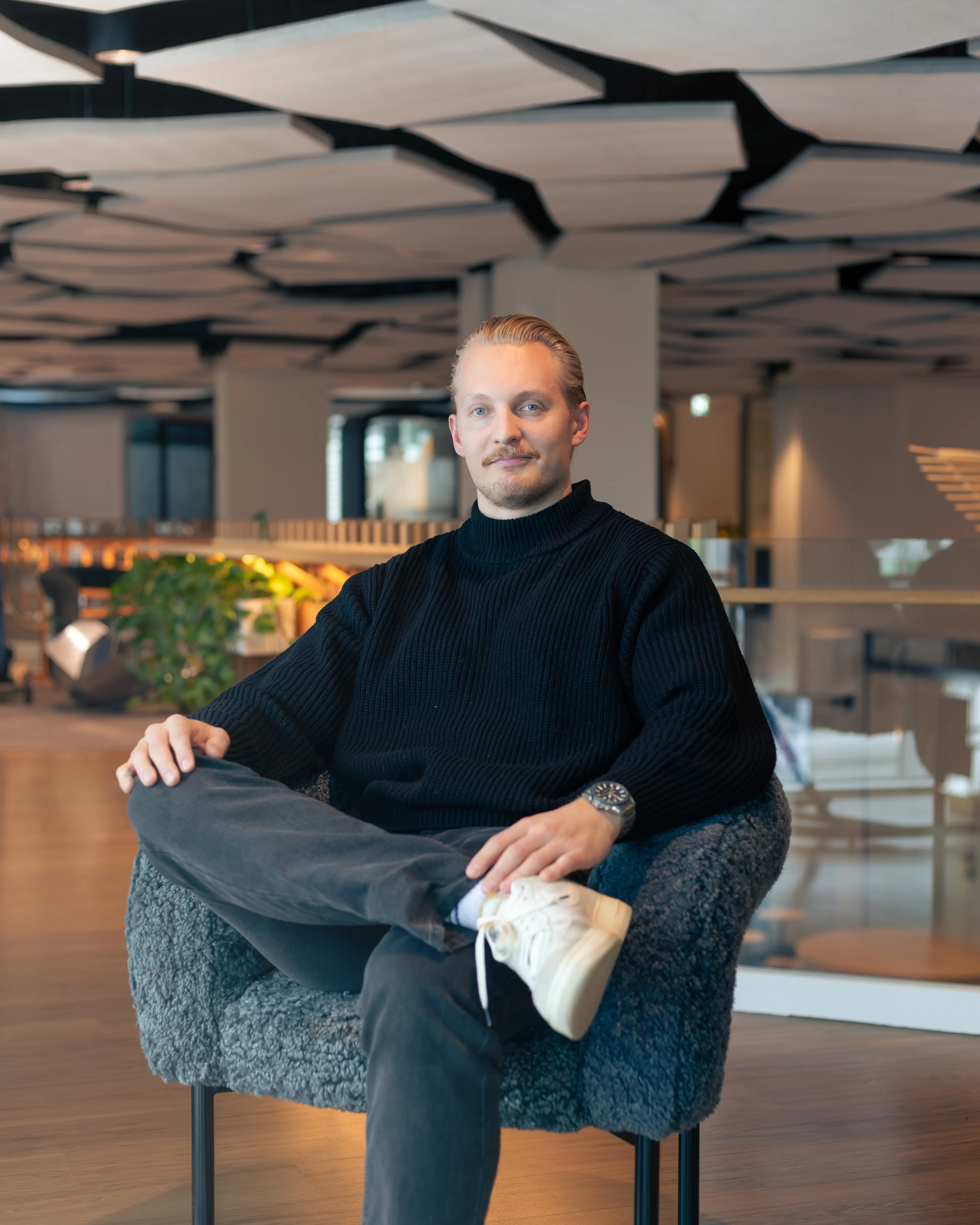 Man with light hair and mustache sitting cross-legged in a modern office space in a black sweater.