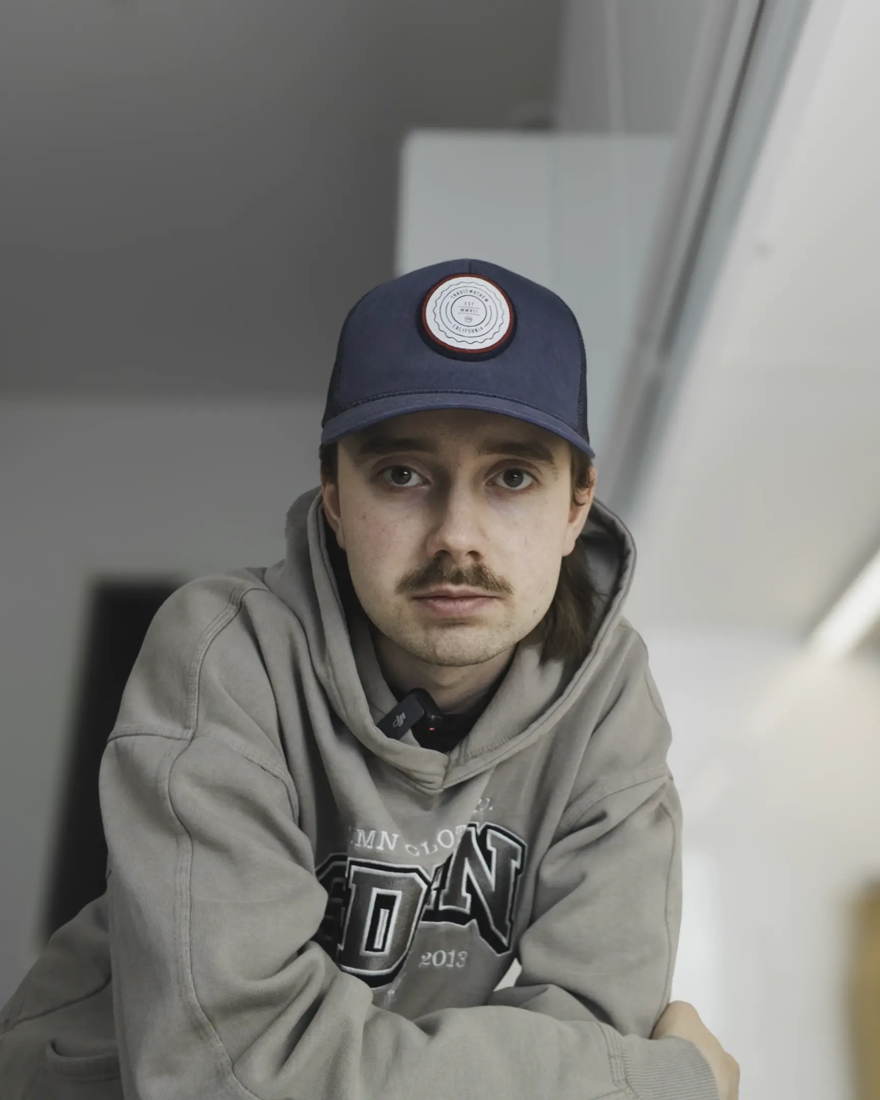 Young man with mustache wearing a blue cap and gray hoodie looking directly at the camera indoors.