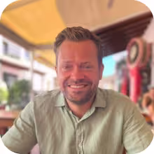 Smiling man with short brown hair and beard wearing a light green shirt, sitting at an outdoor cafe.