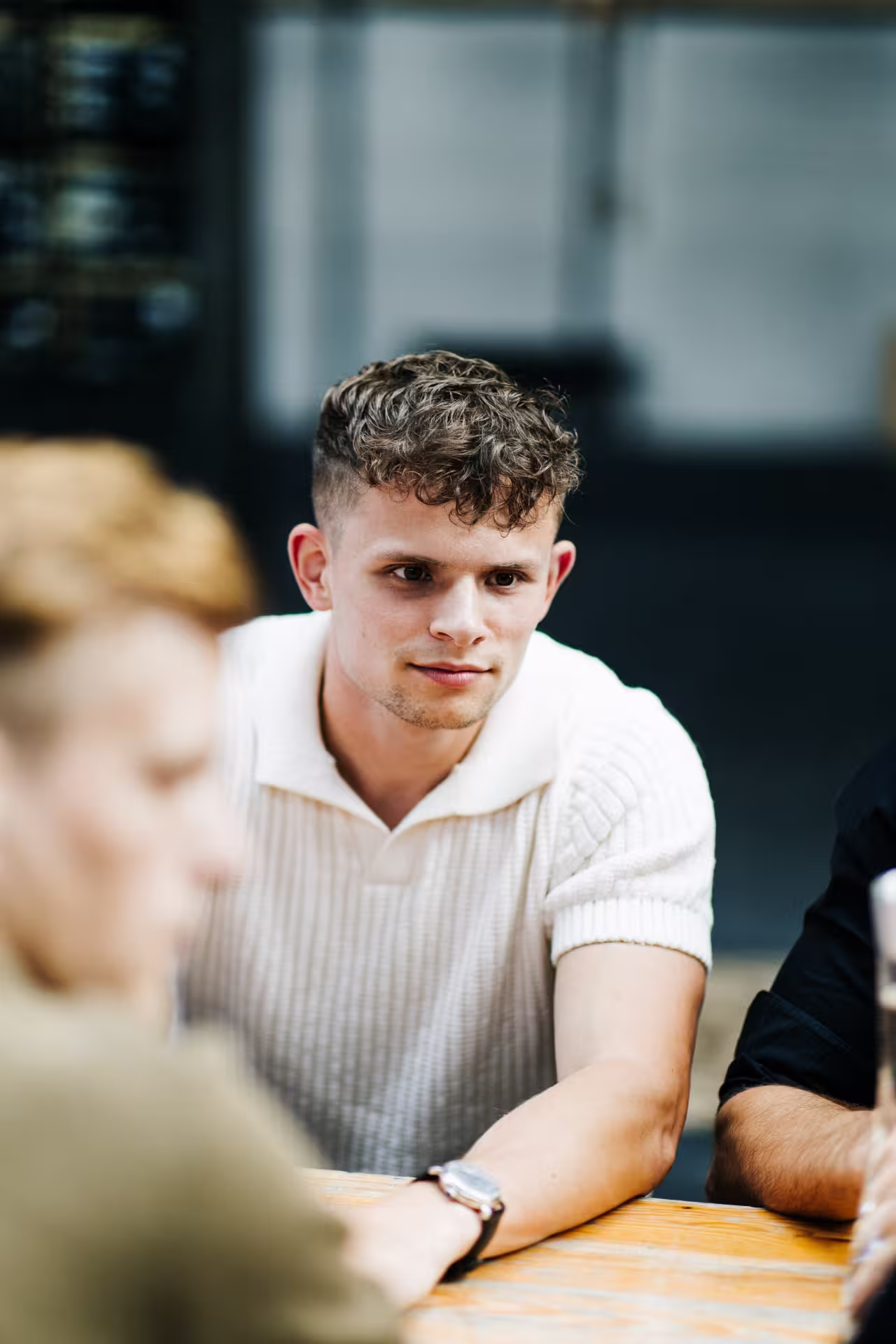 Young man with curly hair wearing a white short-sleeve sweater sitting at a table, looking attentively forward.