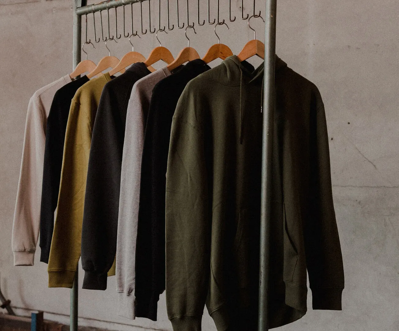 A selection of hooded sweatshirts in various colors including pink, yellow, gray, and dark green, hanging on wooden hangers from a metal rack. The hoodies are displayed against a neutral, textured background, emphasizing their casual style and the variety of colors available.