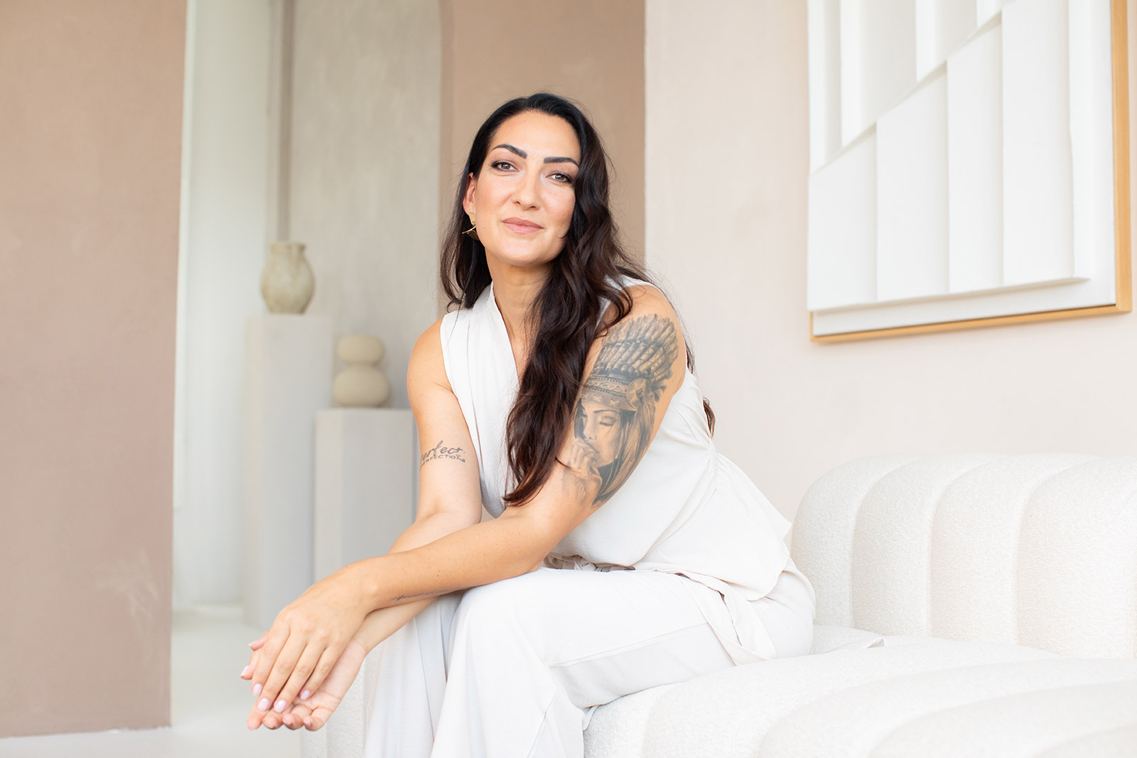 Frau mit langen dunklen Haaren, die auf einem hellen Sofa sitzt und ein weißes Outfit trägt, mit einem auffälligen Tattoo auf dem linken Oberarm.