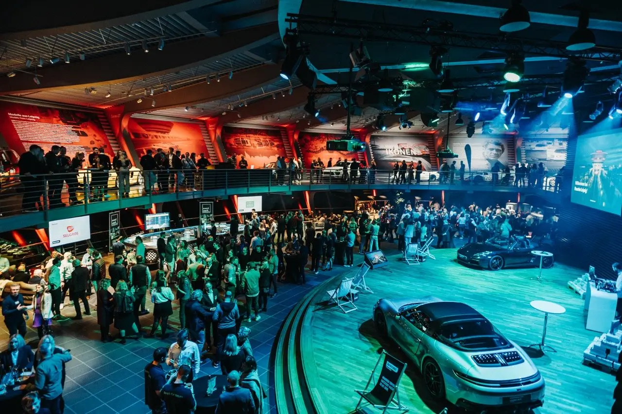Große Menschenmenge in einer modernen Eventhalle mit zwei Ebenen. Unten stehen Besucher in Gruppen zusammen, während oben weitere Gäste entlang einer Galerie laufen. Im Vordergrund sind mehrere ausgestellte Sportwagen auf einer beleuchteten Fläche zu sehen