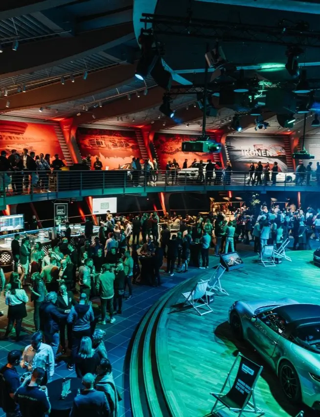 Große Menschenmenge in einer modernen Eventhalle mit zwei Ebenen. Unten stehen Besucher in Gruppen zusammen, während oben weitere Gäste entlang einer Galerie laufen. Im Vordergrund sind mehrere ausgestellte Sportwagen auf einer beleuchteten Fläche zu sehen