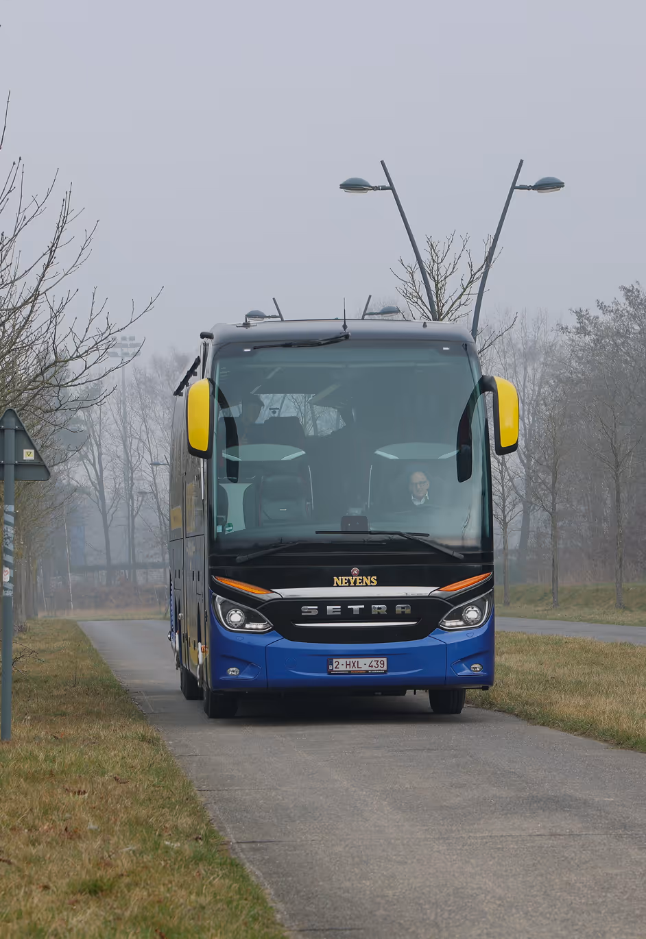 Vooraanzicht van een blauwe en zwarte touringcar met gele spiegels van Neyens met Marc hoeks als chauffeur op een lege straat met bomen in mistige omgeving.