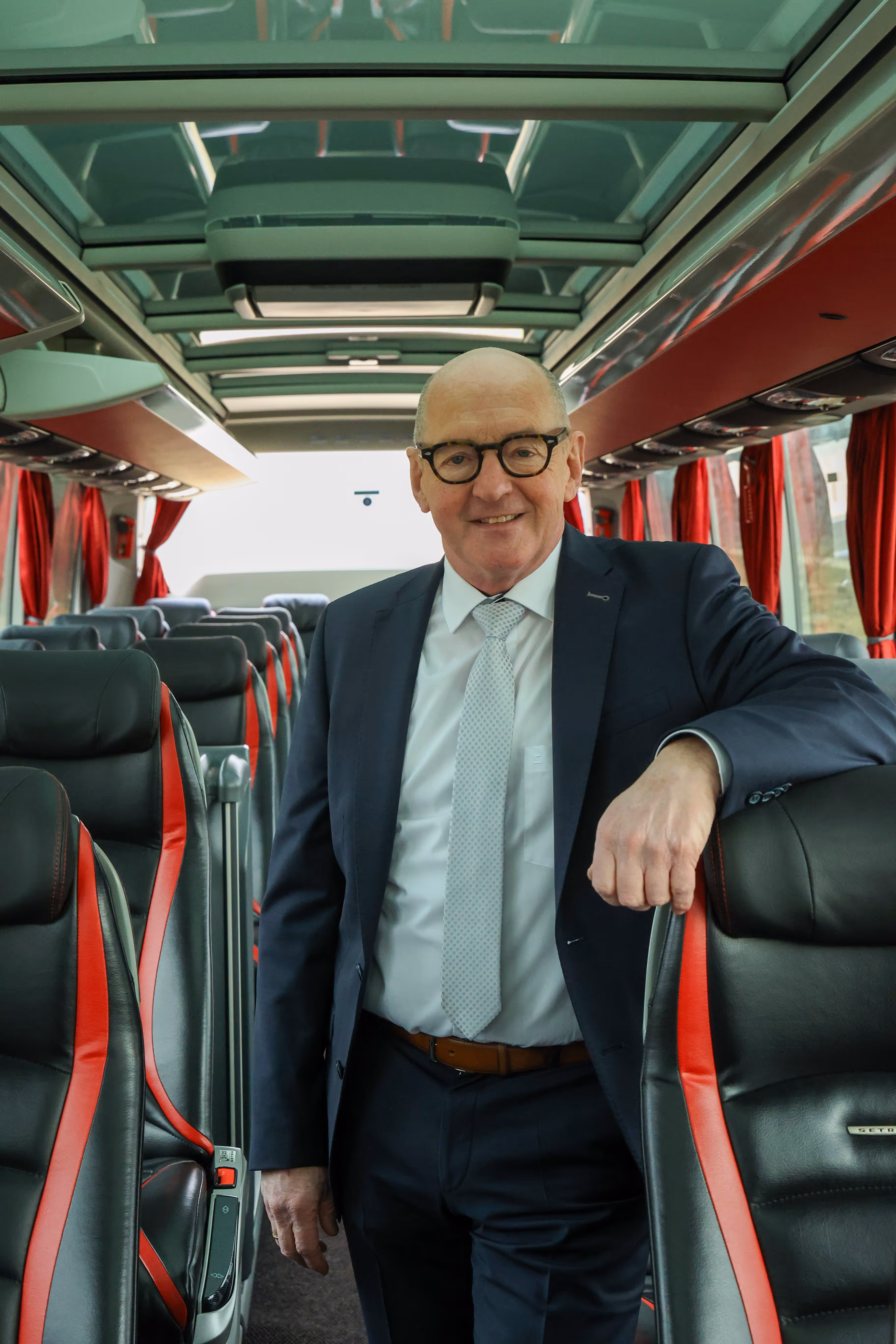 Marc in pak met bril en stropdas leunt ontspannen op de leuning van een busstoel in een lege luxe bus.